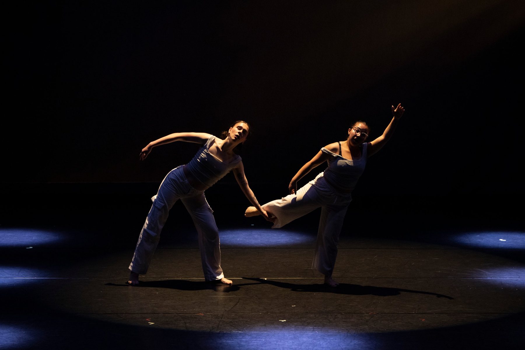 Two dancers in white lean in opposite directions on a dark stage, one with a leg lifted, lit by pools of blue light.