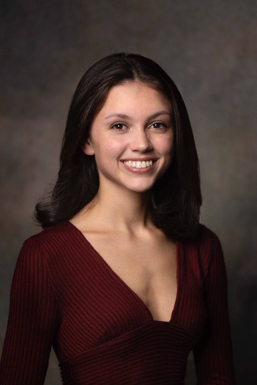 Daniella Molinaro smiling, with shoulder-length dark hair and a burgundy ribbed top, against a dark gray backdrop.