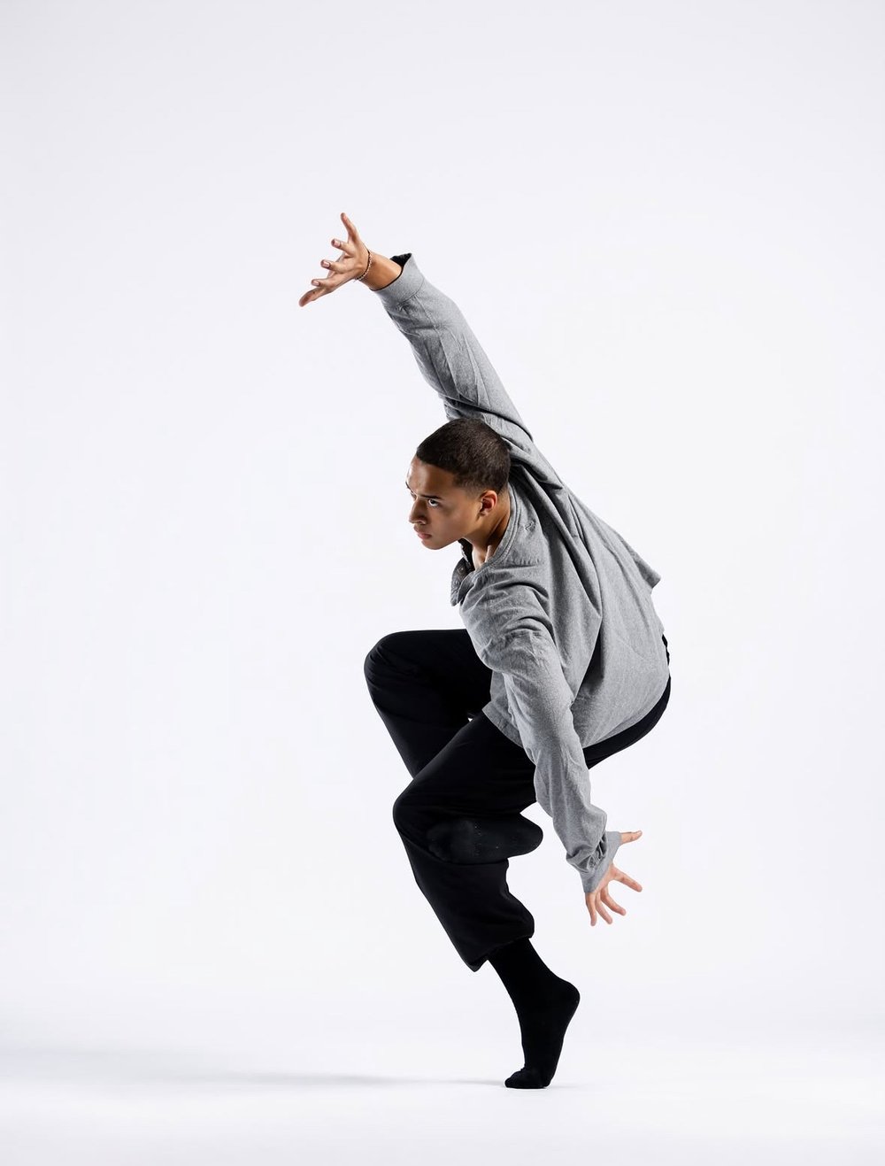 David McCool mid-dance against a white backdrop in a gray sweatshirt and black pants, one arm stretched high and one knee lifted.