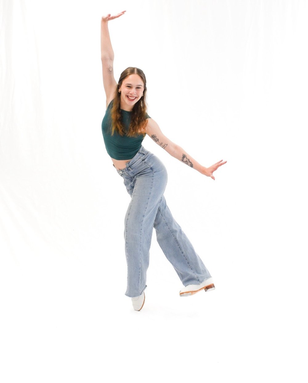 Kelsie Shinall mid-dance in tap shoes, one arm raised high and the other stretched out, against a white backdrop.