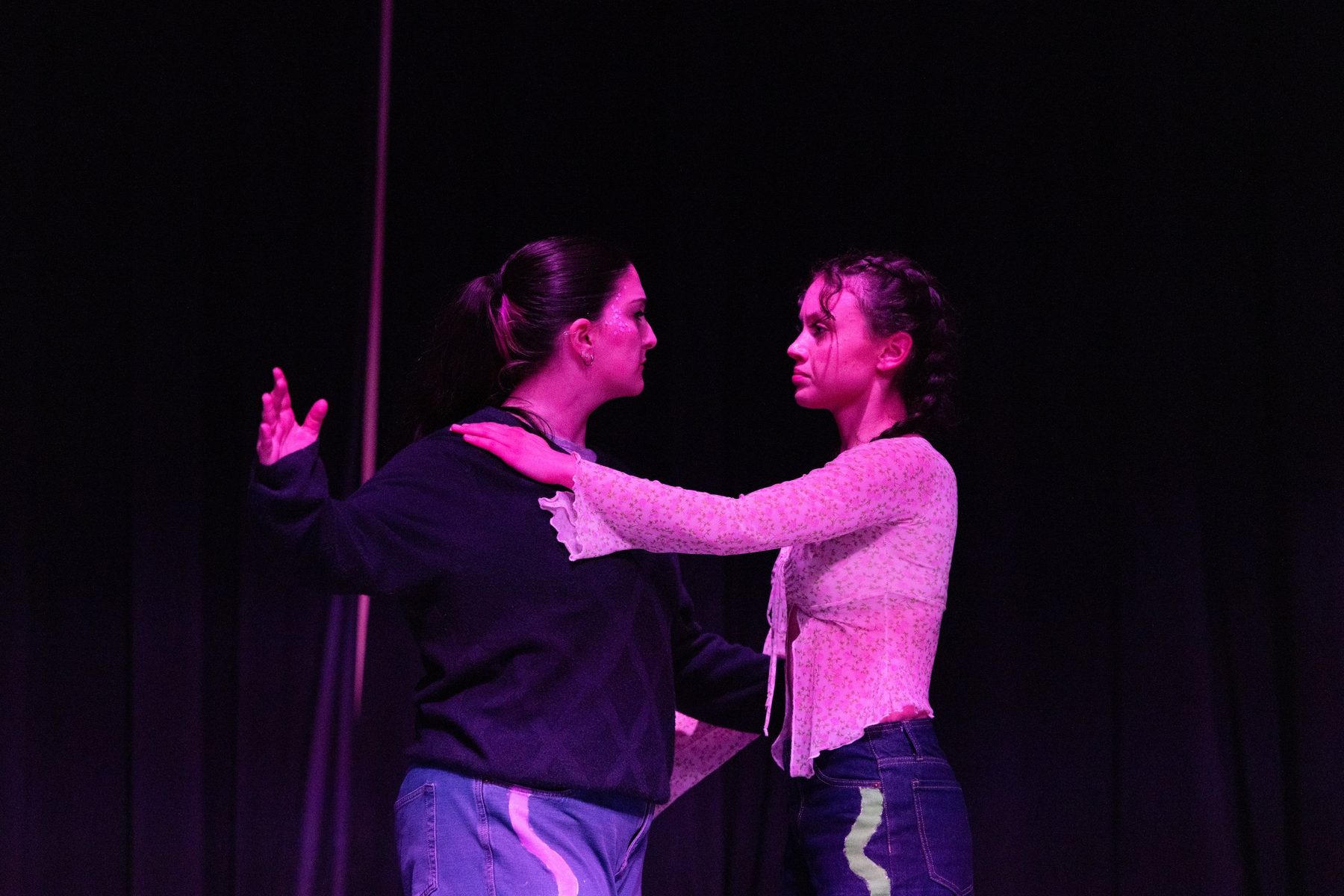 Two dancers face each other in magenta stage light, one resting a hand on the other's shoulder.