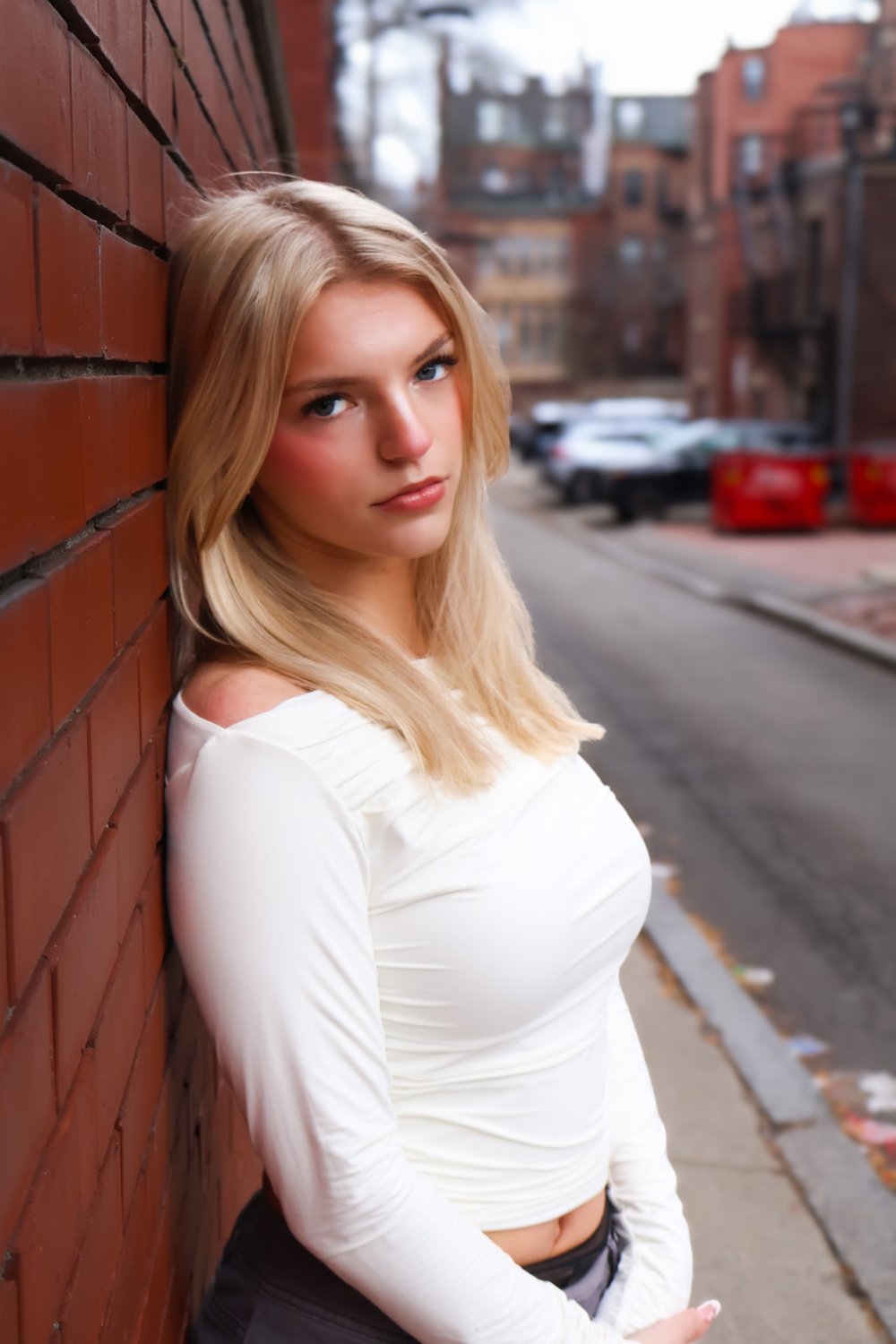 Summer Lee Edwards leaning against a red brick wall on a city street, wearing a white long-sleeve top.